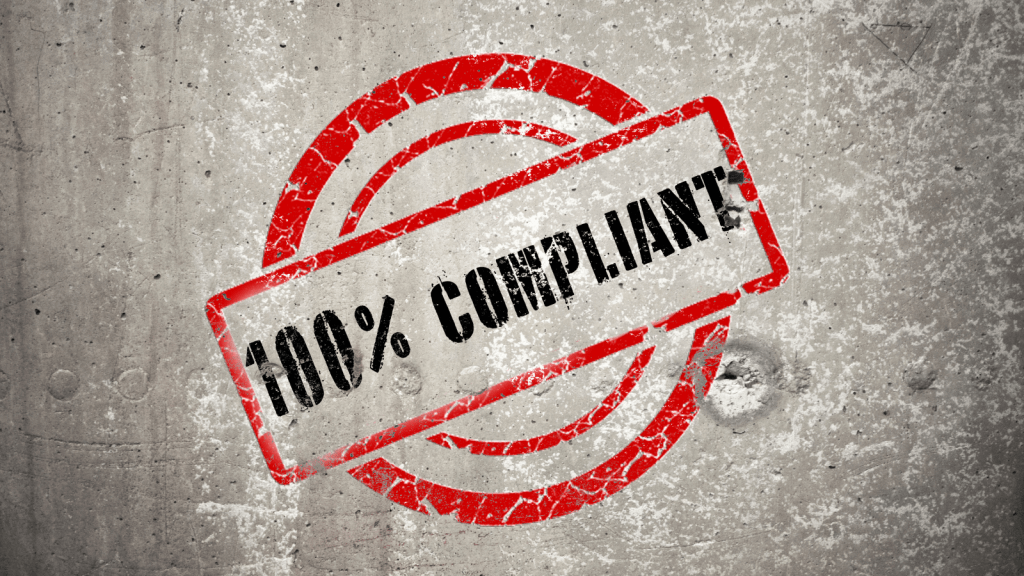 Image of a red circular stamp with the bold black text “100% COMPLIANT” printed diagonally across it on a gray, textured concrete background. The design conveys full compliance or certification.