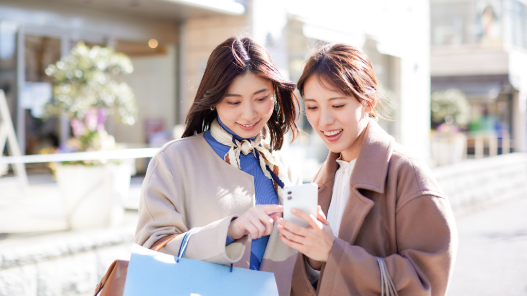 Two women standing outdoors smiling and looking at a smartphone together, one holding a shopping bag, representing social connection and digital engagement in a modern lifestyle setting.
