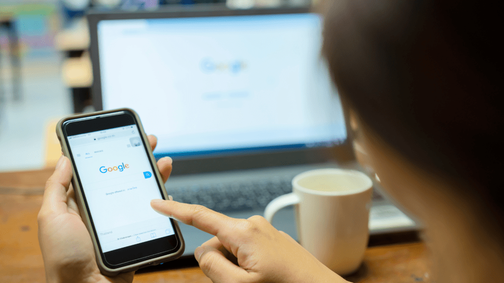 Person holding a smartphone and tapping the Google search page while a laptop with the Google homepage is open in the background on a desk with a coffee mug.